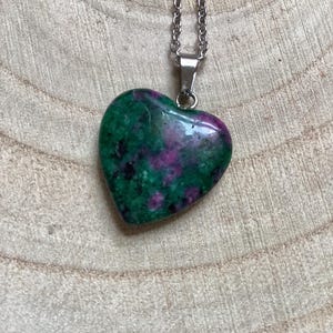 Könnte beinhalten: Halskette mit herzförmigem Anhänger aus poliertem Stein in Grün und Rosa. Der Anhänger hängt an einer silberfarbenen Kette. Die Halskette wird auf einer Holzoberfläche präsentiert.
