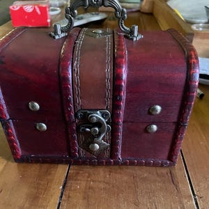 May include: A small, antique-style wooden chest with a dark red finish. It features a brown leather strap and handle, along with decorative metal hardware and a clasp. The chest is adorned with brass-colored studs.