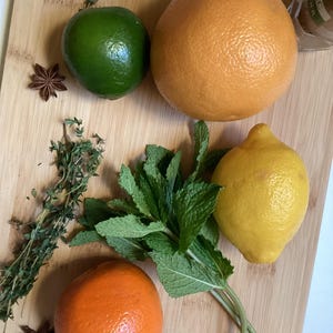 以下が含まれることがあります： 柑橘系の飲み物を作るための材料が置かれた木製のまな板。ライム、グレープフルーツ、レモン、ミントの葉、タイムの葉、スターアニスなどがあります。