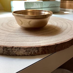 Könnte beinhalten: Eine kleine, goldfarbene Schale mit einem Design aus dreifachem Mond und Pentakel. Die Schale steht auf einer runden Holzscheibe mit natürlichem Rindenrand. Die Schale hat eine glatte, polierte Oberfläche.