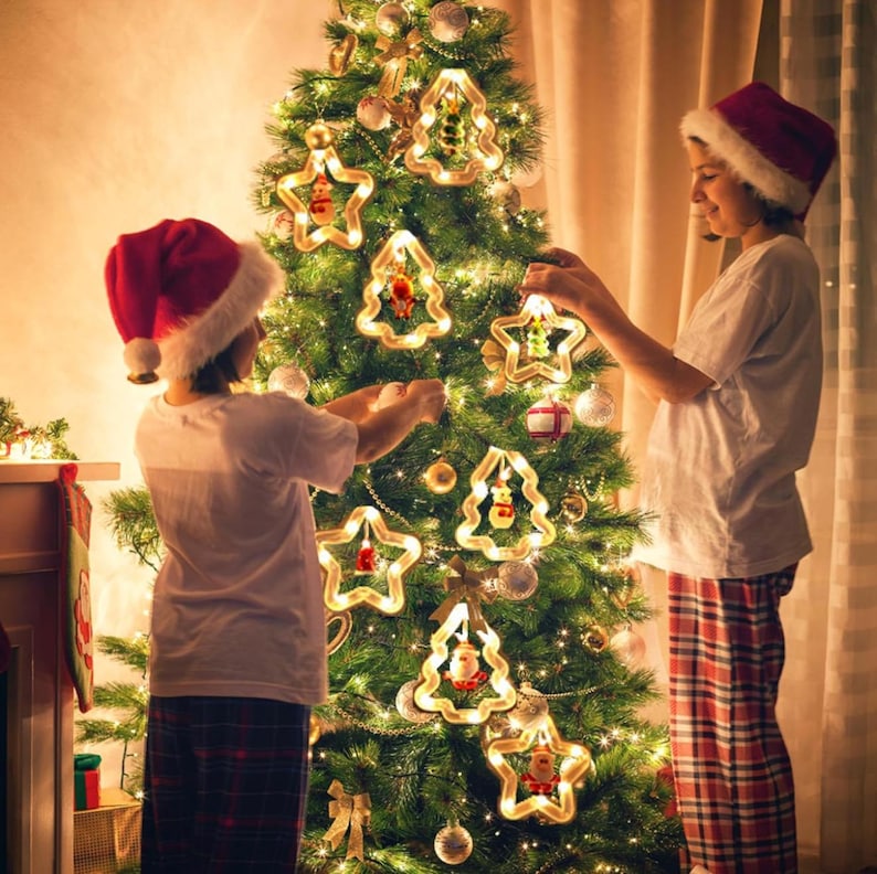 May include: Two children decorate a Christmas tree with string lights and star and tree shaped ornaments. The tree is decorated with white string lights and has a variety of ornaments including stars, trees, and other shapes. The children are wearing red and white pajamas.