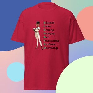 May include: Red t-shirt with a black and white illustration of a woman holding a book. The text "ELEVATED" is written vertically in green, with each letter representing a word: Educated, Ladies, Evolving, Vitalizing, And, Transcending, Excellence, Determinedly.