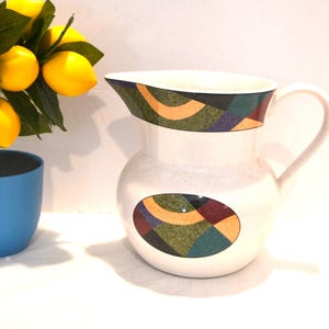 May include: White ceramic pitcher with a curved handle and a spout. The pitcher features a colorful geometric design around the rim and on an oval shape on the body. A small lemon tree in a blue pot is in the background.