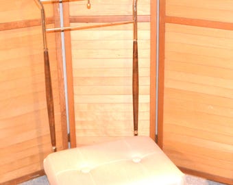 May include: A vintage valet stand with a cream-colored padded seat and a metal frame. The stand has a coat hanger, a bar for trousers, and wooden accents. The background includes a wooden folding screen.