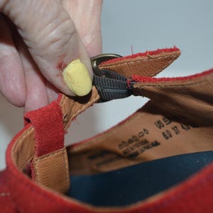 May include: Close-up of a red suede sandal with a buckle closure. The sandal has a brown leather insole and a black strap. The size is US 6.0, EU 37, and UK 4.0.