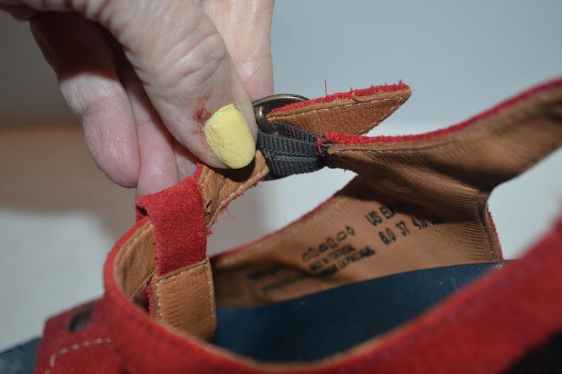 May include: Close-up of a red suede sandal with a buckle closure. The sandal has a brown leather insole and a black strap. The size is US 6.0, EU 37, and UK 4.0.