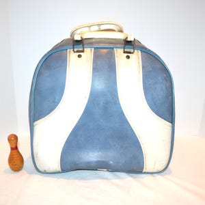 Könnte beinhalten: Vintage Bowlingtasche in Blau und Weiß mit weißem Griff und Metallverschlüssen. Neben der Tasche befindet sich eine Holz-Bowlingkegel mit roten Streifen. Die Tasche hat eine abgerundete Form und ein Retro-Design.