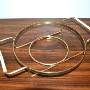 Op de afbeelding: Een goudkleurige metalen onderzetter met een cirkelvormig ontwerp en een handvat met een witte accent. De onderzetter heeft een kleine binnenste cirkel en is ontworpen om oppervlakken te beschermen tegen hete gerechten. De onderzetter staat op een donker houten oppervlak.