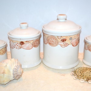 May include: A set of four white ceramic canisters with lids, decorated with a band of seashells in shades of brown and tan. A large seashell and a small woven mat with shells are also present, suggesting a coastal theme.