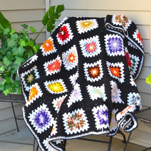 Puede incluir: Una manta de crochet en blanco y negro con cuadrados coloridos. La manta está hecha de cuadrados de la abuela en varios colores, incluyendo rojo, naranja, amarillo, verde, azul, morado y blanco. La manta está doblada y drapeada sobre una silla.