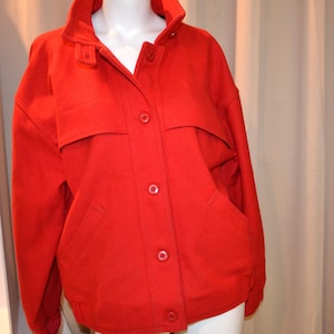 May include: A bright red jacket with a high collar and button-up front. The jacket has two front pockets and a flap detail across the chest. The jacket is displayed on a mannequin.