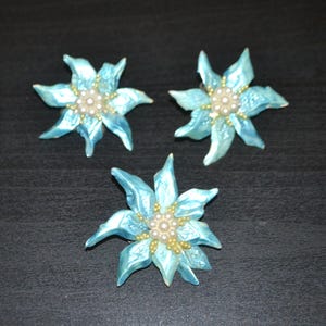 May include: Three light blue flower-shaped brooches with pearl and gold bead centers. The brooches have pointed petals and are arranged on a dark surface. These decorative brooches could be used as accessories.