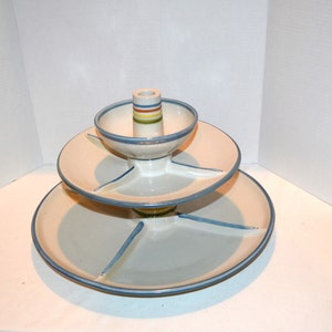Puede incluir: Un plato de servir de cerámica de tres niveles con un pedestal central y cuatro secciones divididas en cada nivel. El plato está decorado con rayas azules y verdes sobre un fondo blanco.