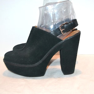 May include: Black platform clogs with a suede-like texture. The shoes have a thick platform sole and a high, block heel. A black strap with a gold-colored buckle secures the ankle. The clogs have a closed toe design.