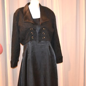 May include: A black dress and matching bolero jacket set. The dress has a flared skirt and the jacket features a notched collar and decorative gold-colored buttons. The outfit is displayed on a mannequin against a neutral background.