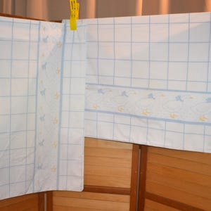 May include: White fabric with a blue grid pattern and a decorative border featuring duck illustrations. The fabric is hanging from a clothesline with blue and yellow clothespins. The fabric appears to be a curtain or a similar home textile.
