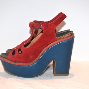 May include: Red suede platform sandal with a blue platform and heel. The sandal features a T-strap design with a buckle closure and a peep-toe front. The platform measures approximately 3 inches.