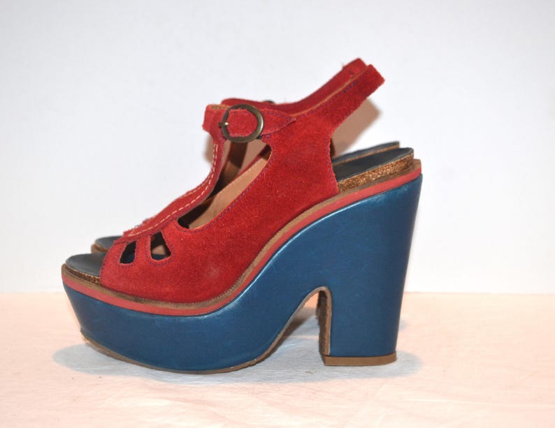 May include: Red suede platform sandal with a blue platform and heel. The sandal features a T-strap design with a buckle closure and a peep-toe front. The platform measures approximately 3 inches.