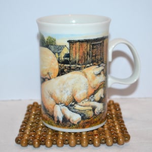 May include: White ceramic mug featuring a pastoral scene with pigs and piglets. The mug has a curved handle and is set on a square wooden bead coaster. The artwork depicts a farm setting with a barn and a blue sky.