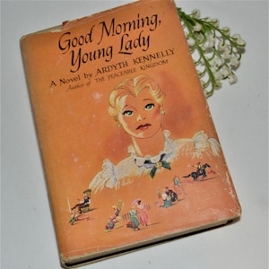 May include: A vintage book cover with an orange background. The title is "Good Morning, Young Lady" by Ardyth Kennelly. The cover features an illustration of a young woman with blonde hair and a white dress.
