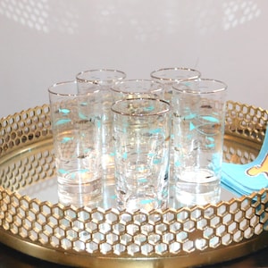 Op de afbeelding: Zes heldere highballglazen met gouden randen en een turquoise visontwerp zijn gerangschikt op een goudkleurig dienblad met honingraatpatroon. Een lichtblauwe servet met een gouden ontwerp ligt op de dienblad.