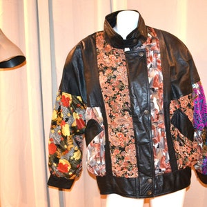 May include: A black leather jacket with a patchwork design. The jacket has a stand-up collar, a zipper closure, and two pockets. The patchwork design features floral patterns in shades of orange, red, purple, and green.