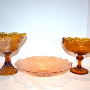 May include: Three decorative glass serving dishes. Two amber-colored pedestal bowls with scalloped rims and one shallow, light-pink, woven-patterned bowl. The pedestal bowls have ornate detailing and are suitable for serving fruit or desserts.