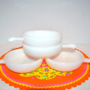 Puede incluir: Tres cuencos de vidrio lechoso blanco con un solo asa, apilados en formación piramidal. Cada cuenco presenta un diseño liso y redondeado con un asa corta que sobresale por un lado. Los cuencos son opacos, brillantes y de color uniforme.