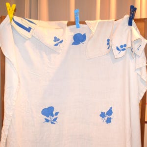 Puede incluir: Ropa de lino blanco con diseños florales azules, colgada en un estante de madera con pinzas. La ropa incluye una camisa y varios pañuelos. Los diseños azules son de flores y hojas.