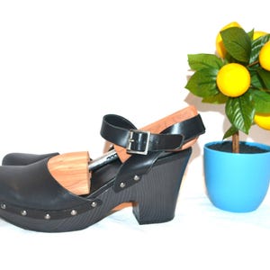 Pode incluir: Tamancos de couro preto com tira de fivela e salto de madeira. Os tamancos têm bico arredondado e são cravejados com rebites prateados. Uma pequena árvore de limão em um vaso azul está ao fundo.