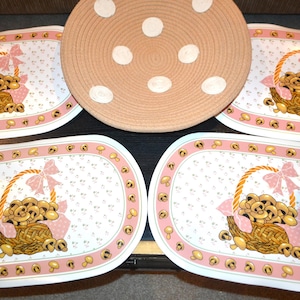 May include: A set of four oval placemats and a round serving mat. The placemats feature a basket of mushrooms with a pink bow and border. The serving mat is tan with white polka dots.