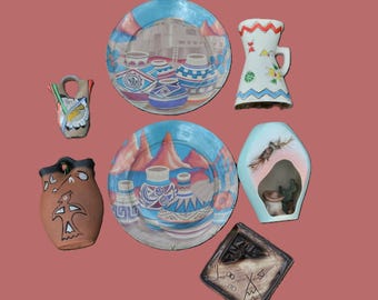 Peut inclure: Une collection d'objets en céramique comprenant deux assiettes rondes avec des paysages désertiques, une applique murale en terre cuite avec un motif d'oiseau, un vase blanc à motifs géométriques, une applique bleue et un plateau carré brun.