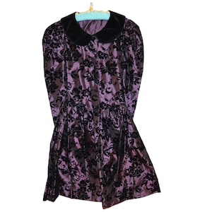 May include: A vintage purple dress with a black floral pattern and a black velvet collar. The dress has long sleeves and a gathered waist. It is hanging on a light blue hanger.