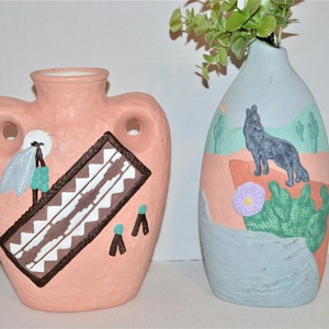 Könnte beinhalten: Zwei Keramikvasen mit handbemalten Designs. Die Vase links ist rosa mit einem braunen und weißen geometrischen Muster, einer Feder und türkisfarbenen Perlen. Die Vase rechts ist hellblau mit einer gemalten Szene eines Kojoten in einer Wüstenlandschaft mit Kakteen und einer lila Blume.