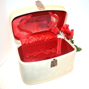 May include: A cream-coloured hard-shell vanity case with a metallic latch and a vibrant red interior lining. The lid features an integrated rectangular mirror framed by red fabric trim, while the main compartment includes a red fabric pocket.