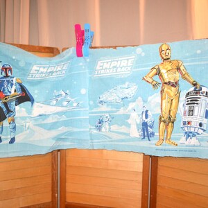 Könnte beinhalten: Ein hellblauer Stoffstreifen mit Grafiken aus Star Wars Das Imperium schlägt zurück. Der Druck zeigt Darth Vader, Boba Fett, C-3PO und R2-D2 neben Raumschiffen und Charakteren in einer Schneelandschaft. Der Stoff wird von bunten Klammern gehalten.