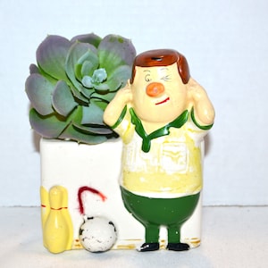 Op de afbeelding: Een keramische plantenpot in de vorm van een cartoonman die een geel shirt en een groene broek draagt. De man houdt zijn handen aan zijn oren en heeft een knipogende uitdrukking. De pot heeft een witte achtergrond met een rood, geel en wit bowlingpin-ontwerp. Een groene succulent plant zit in de pot.