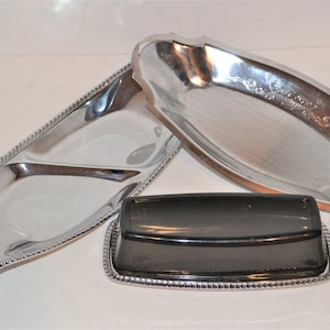 Puede incluir: Tres bandejas de servir plateadas con bordes perlados. La bandeja más grande es de forma ovalada y tiene un diseño decorativo. La bandeja del medio es rectangular y está dividida en tres secciones. La bandeja más pequeña es cuadrada y tiene una base negra.