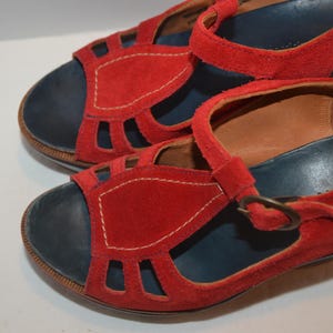 May include: A pair of red suede sandals with a dark blue footbed and a strap with a buckle. The sandals have cut-out details on the vamp and are stitched with white thread.
