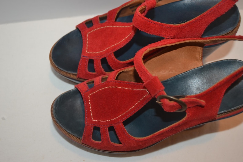 May include: A pair of red suede sandals with a dark blue footbed and a strap with a buckle. The sandals have cut-out details on the vamp and are stitched with white thread.