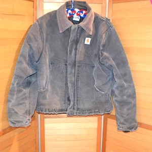 Può includere: Una giacca Carhartt grigio antracite con colletto a punta, chiusura frontale con zip e due ampie tasche inferiori. Il capo presenta una piccola toppa quadrata con logo sul petto sinistro e maniche lunghe con polsini. Il tessuto appare usurato.