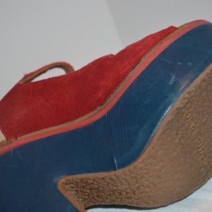 May include: A red suede sandal with a blue platform sole and a brown rubber outsole. The sandal features a strap and a platform heel. The platform sole is approximately 3 inches high.