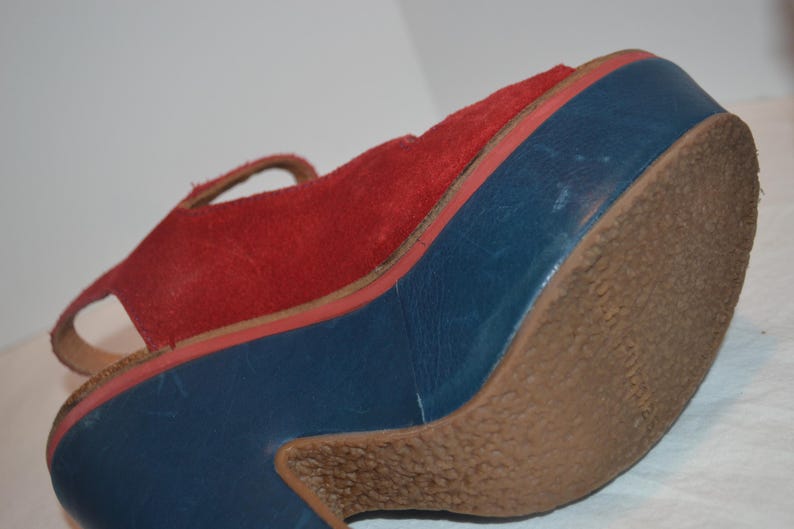 May include: A red suede sandal with a blue platform sole and a brown rubber outsole. The sandal features a strap and a platform heel. The platform sole is approximately 3 inches high.