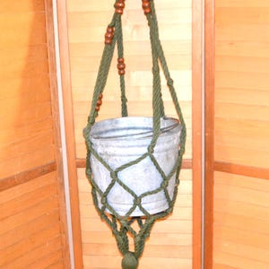 Puede incluir: Un colgador de plantas hecho a mano con un cubo de metal galvanizado. El macetero de macramé es de color verde oliva con cuentas de madera y una borla. El colgador de plantas está suspendido de un anillo de metal azul.