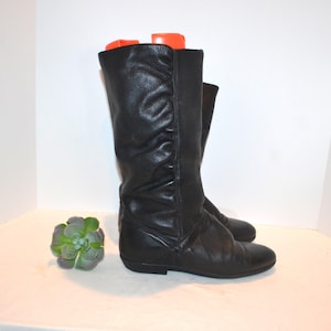 Op de afbeelding: Een paar zwarte leren kniehoge laarzen. De laarzen hebben een ronde neus en een lage hak. Het leer is licht gekreukeld. De laarzen zijn ongeveer 38 cm hoog. De laarzen lijken in goede staat te zijn.