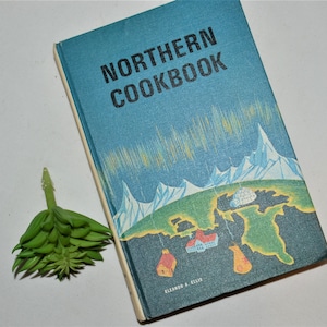 May include: A blue hardcover cookbook titled "Northern Cookbook" with a colorful illustration of a snowy mountain range and a green island with a small village on the cover. The author's name, Eleanor J. Ellis, is printed on the bottom of the cover.