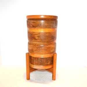 Peut inclure: Un support de plante en bois avec un corps cylindrique et une base à trois pieds. Le support est fait de différentes nuances de bois brun, avec une finition polie. Le design est moderne du milieu du siècle.
