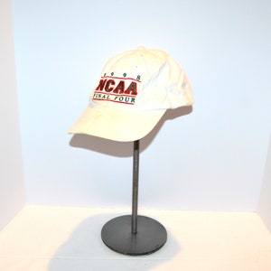 Puede incluir: Gorra de béisbol blanca con el texto "NCAA FINAL FOUR 1998" en rojo y verde. La gorra se muestra sobre un soporte metálico con base circular.