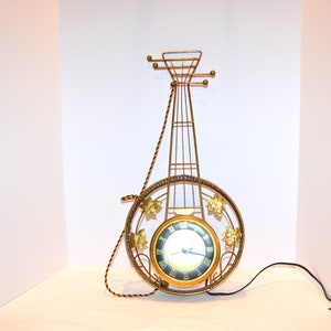 May include: A gold-toned metal wall clock shaped like a banjo with a black face and Roman numeral hour markers.