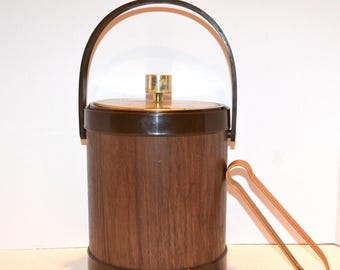 May include: A vintage ice bucket with a dark brown wood grain finish and a matching handle. The lid has a gold-colored center and a brass knob. Copper-colored ice tongs rest beside the bucket. A classic barware item.